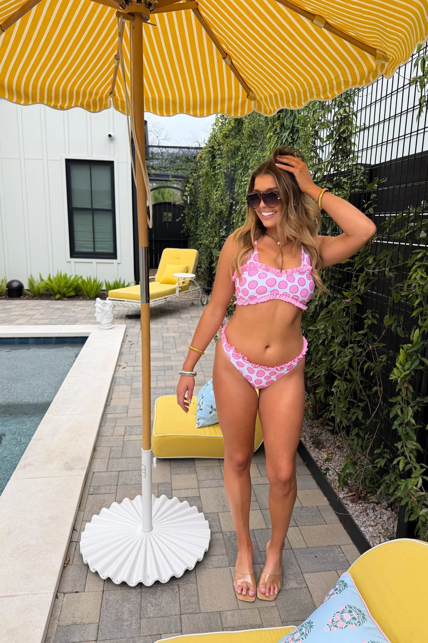 Woman in a pink bikini standing under a yellow striped umbrella by a pool.