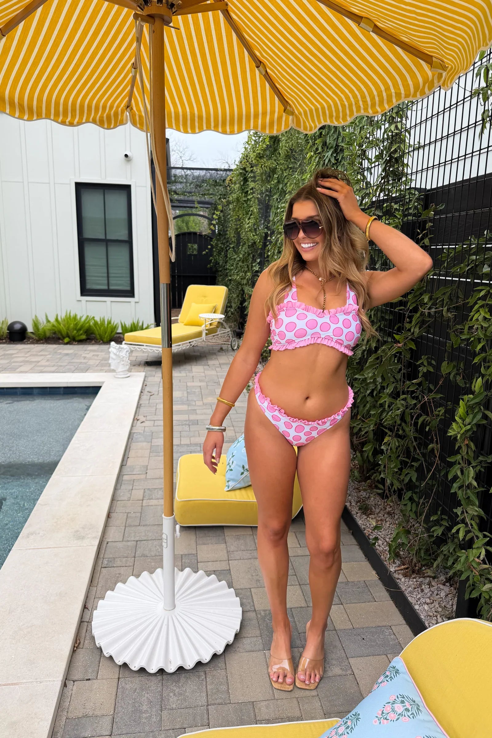 Woman in a pink bikini standing under a yellow striped umbrella by a pool.