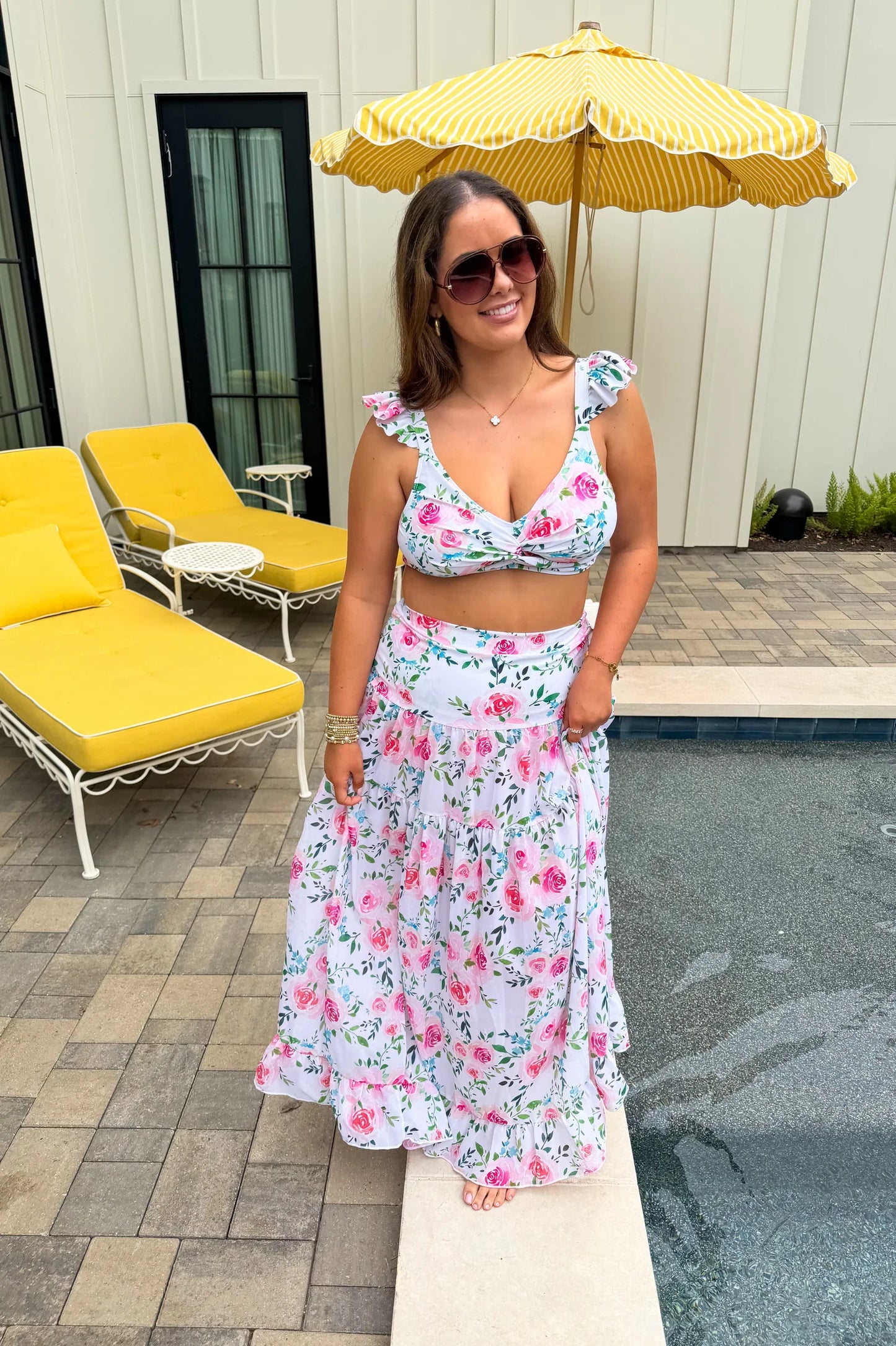Woman in floral dress standing by a pool with yellow umbrella and chairs.