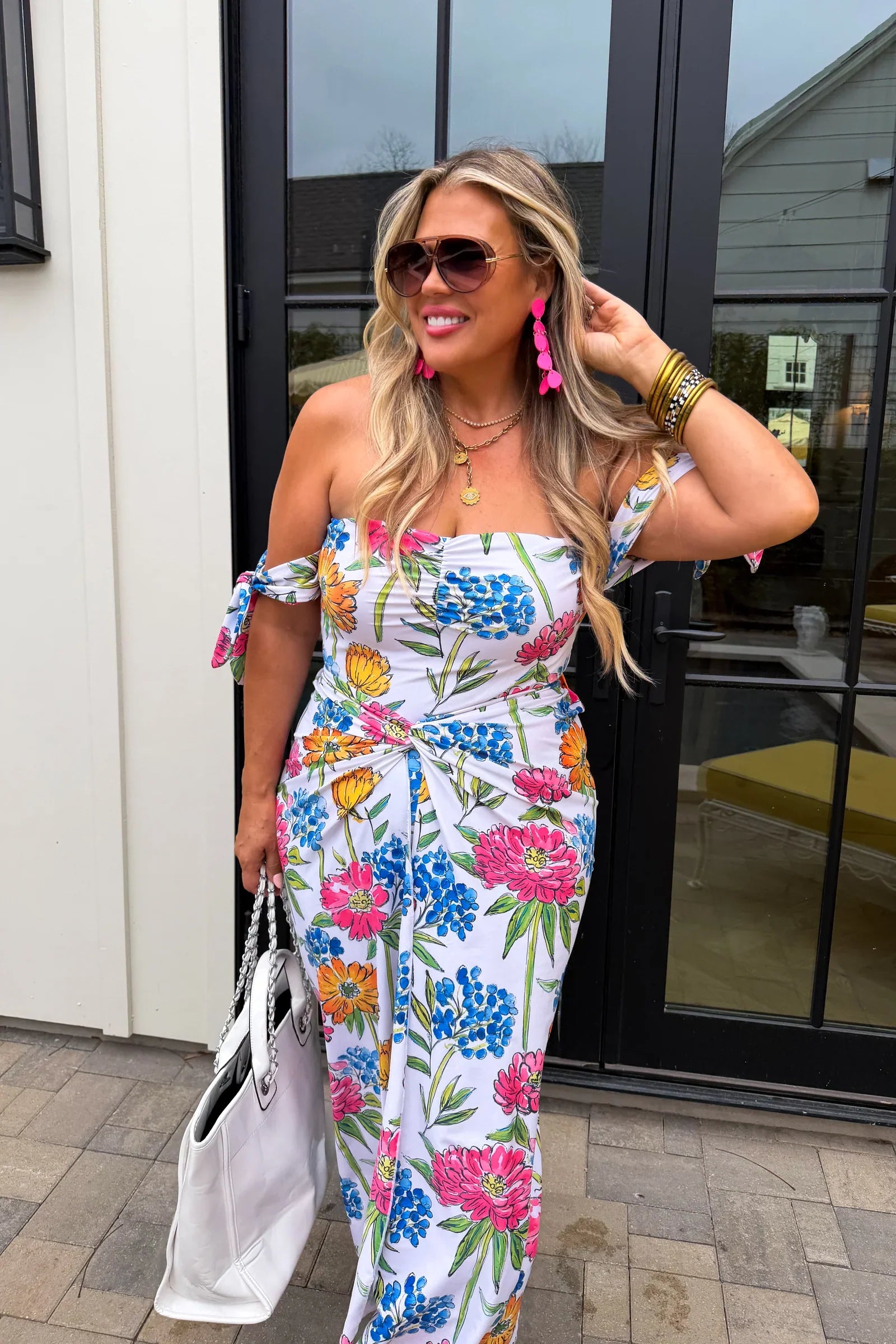 Woman wearing a colorful floral dress with sunglasses and a handbag, standing outside.