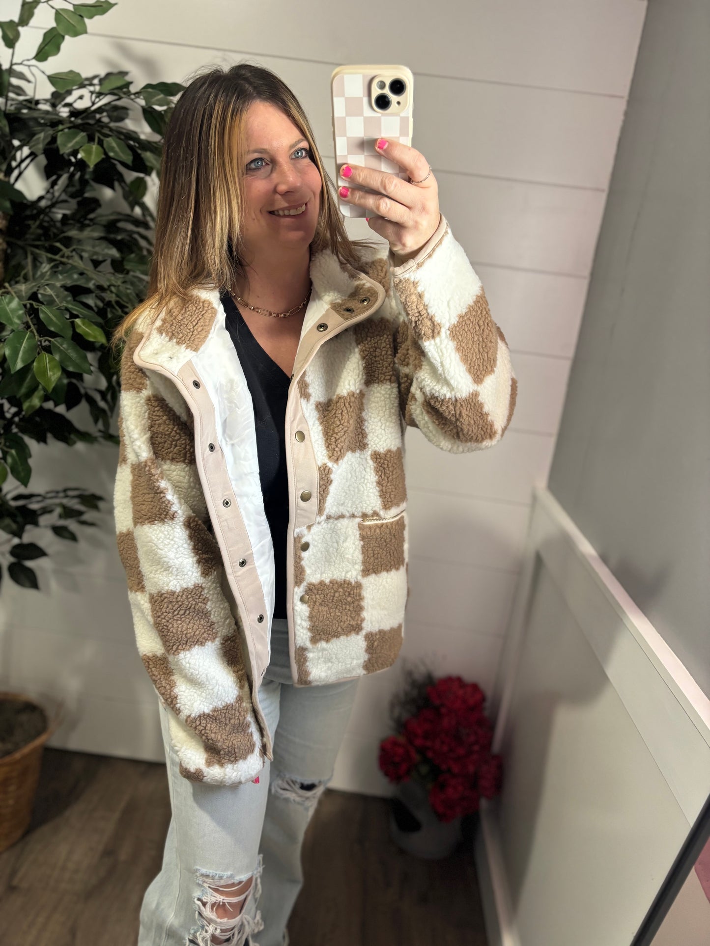 A person wearing a beige and white checkered snap button sherpa jacket, paired with ripped jeans, taking a mirror selfie.
