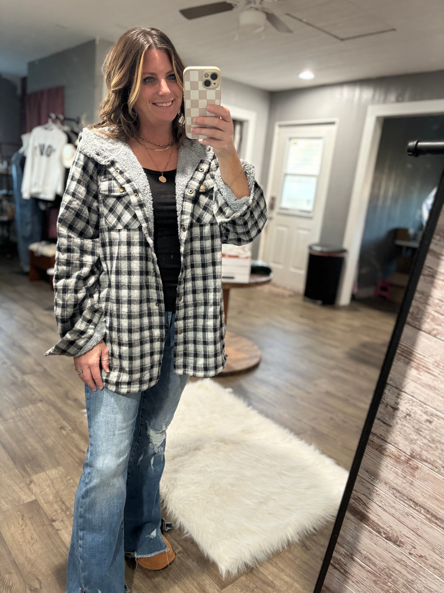 Woman taking a mirror selfie wearing a plaid jacket and jeans in a room with wooden flooring.