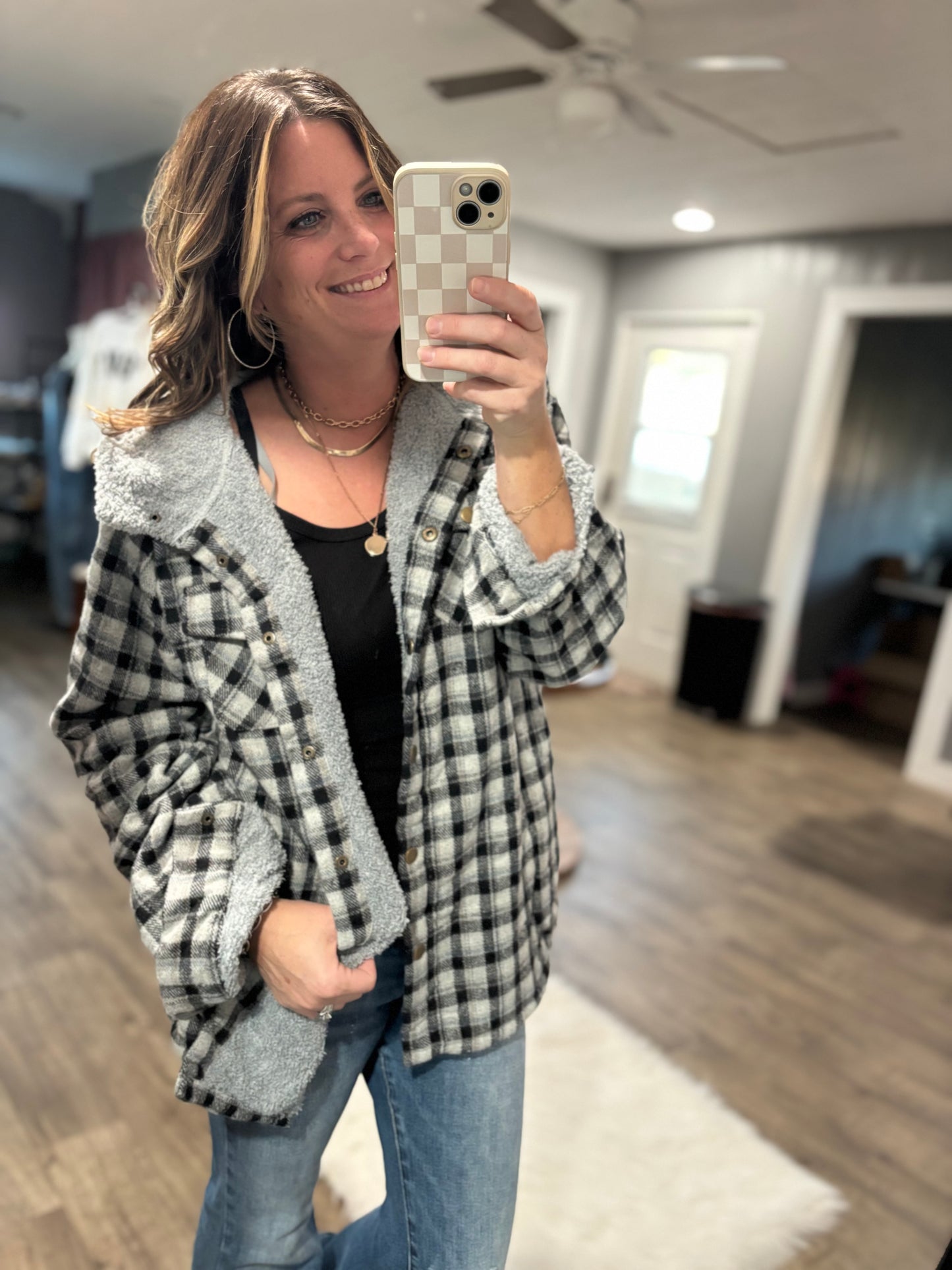 Woman taking a mirror selfie wearing a plaid jacket in a room with wooden flooring and a ceiling fan.