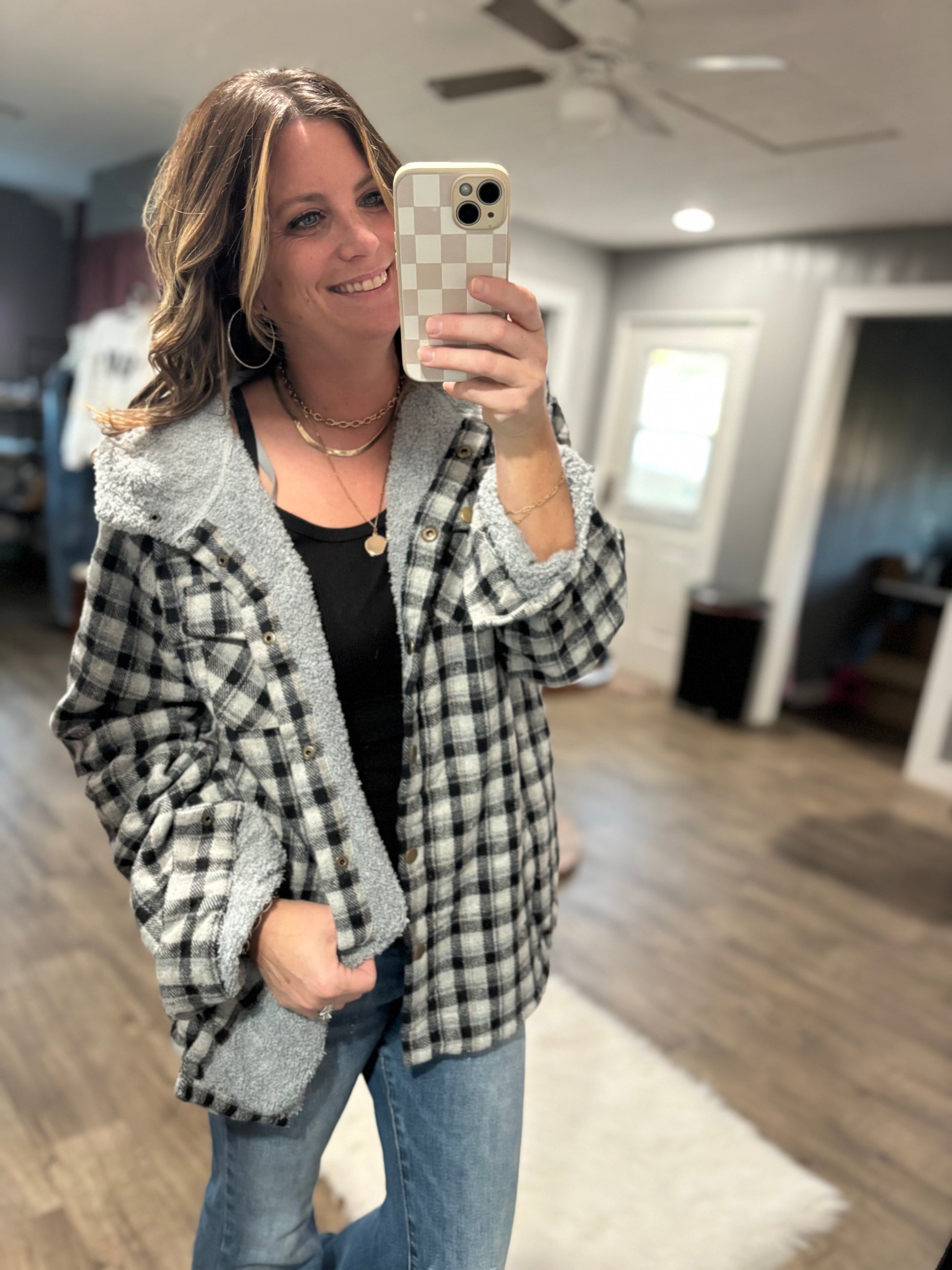 Woman taking a mirror selfie wearing a plaid jacket in a room with wooden flooring and a ceiling fan.