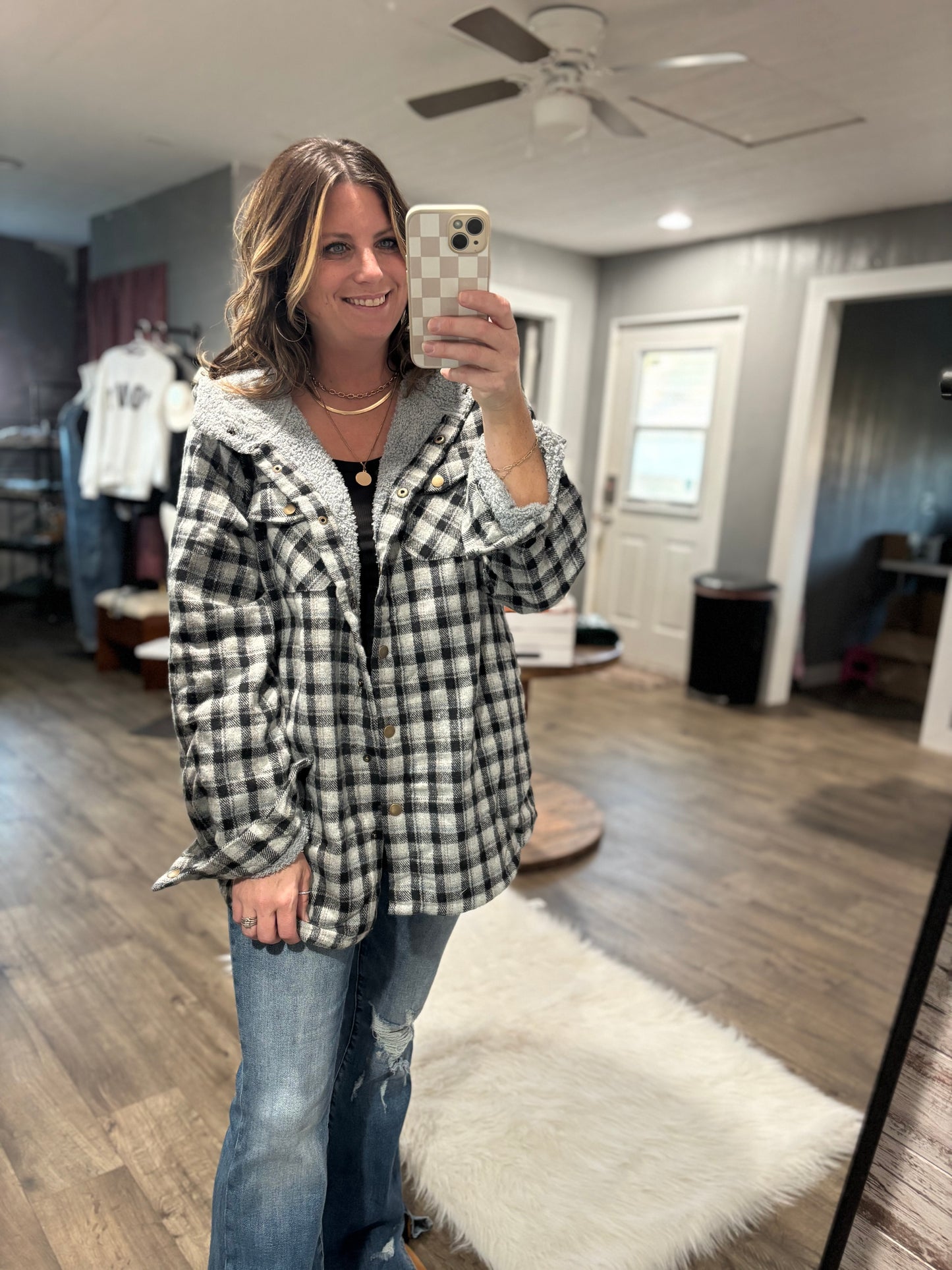 Person taking a mirror selfie wearing a plaid shirt and jeans in a room with wooden flooring and a ceiling fan.