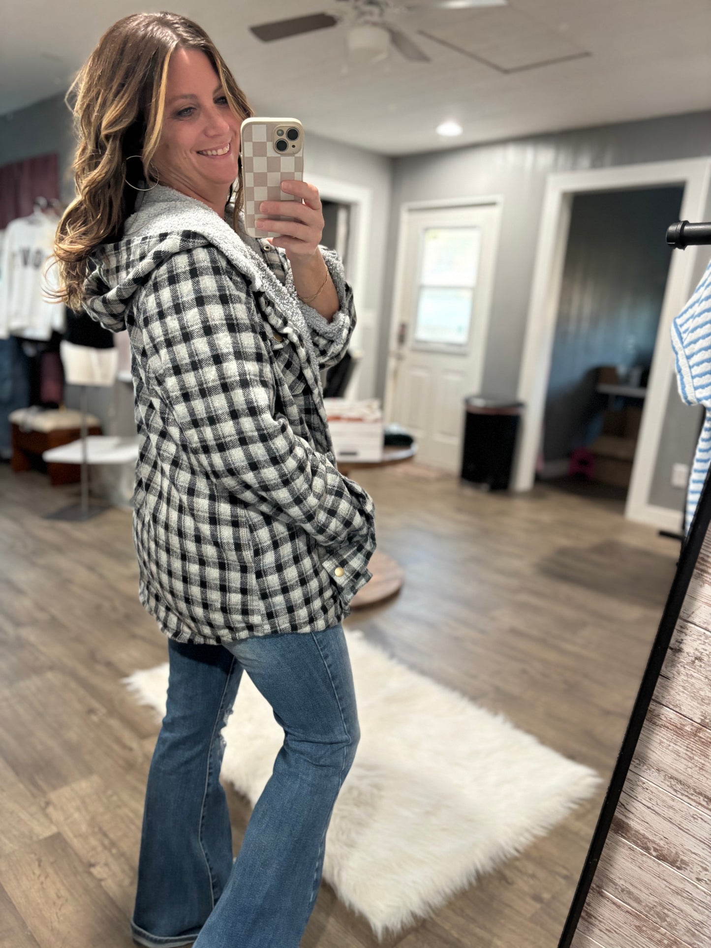 Woman taking a mirror selfie in a living room wearing a plaid jacket and jeans.