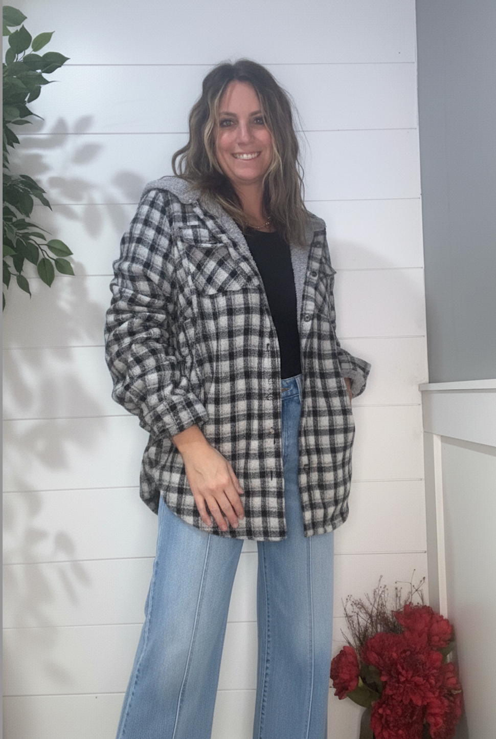 Woman wearing a plaid jacket and jeans standing against a white wall with a plant and flowers in the background.