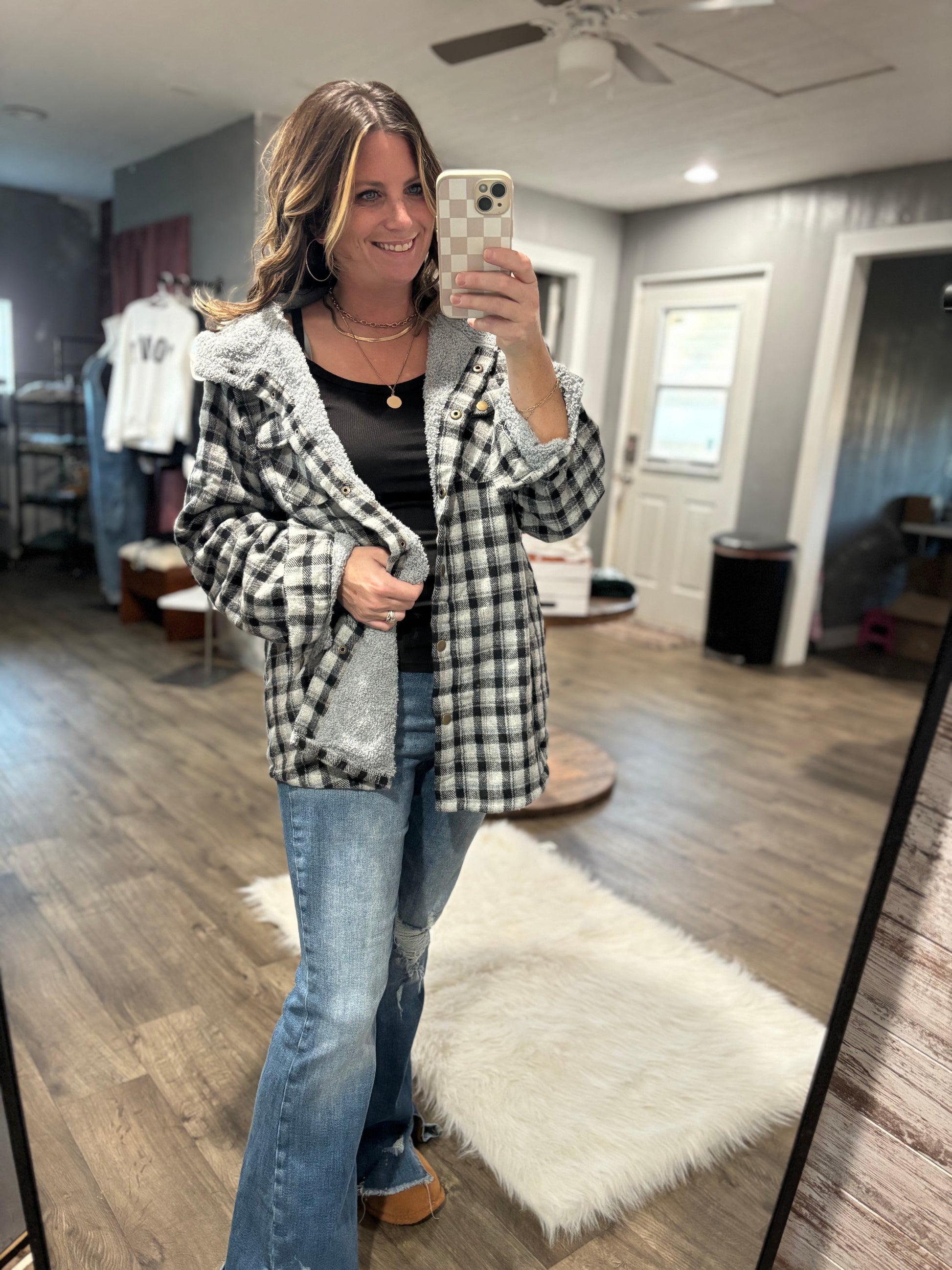Woman taking a mirror selfie wearing a checkered jacket and jeans in a room with wooden flooring.