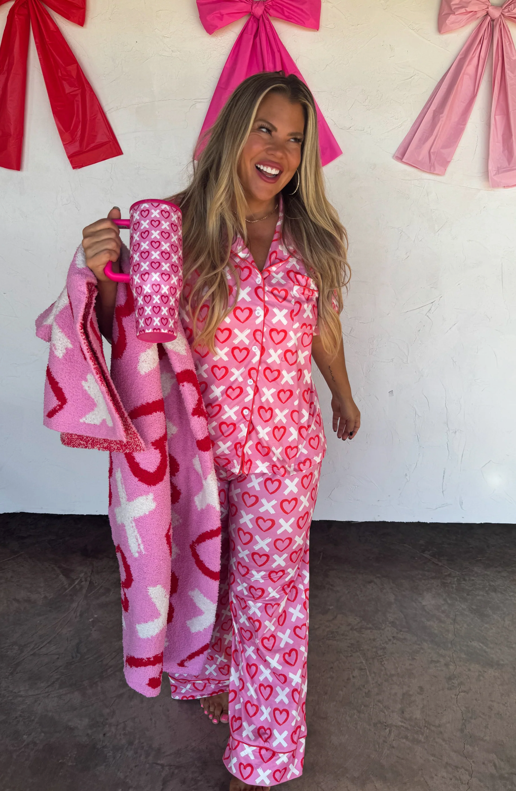 Woman in pink heart-patterned pajama set holding a matching blanket with decorative bows in the background.