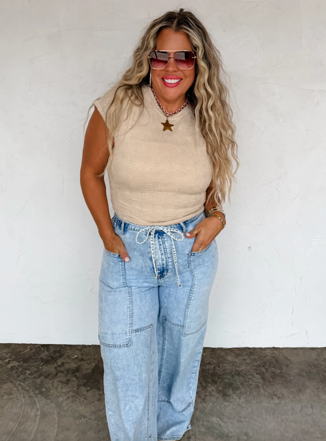 Woman wearing a beige sleeveless top and light blue jeans against a white wall.