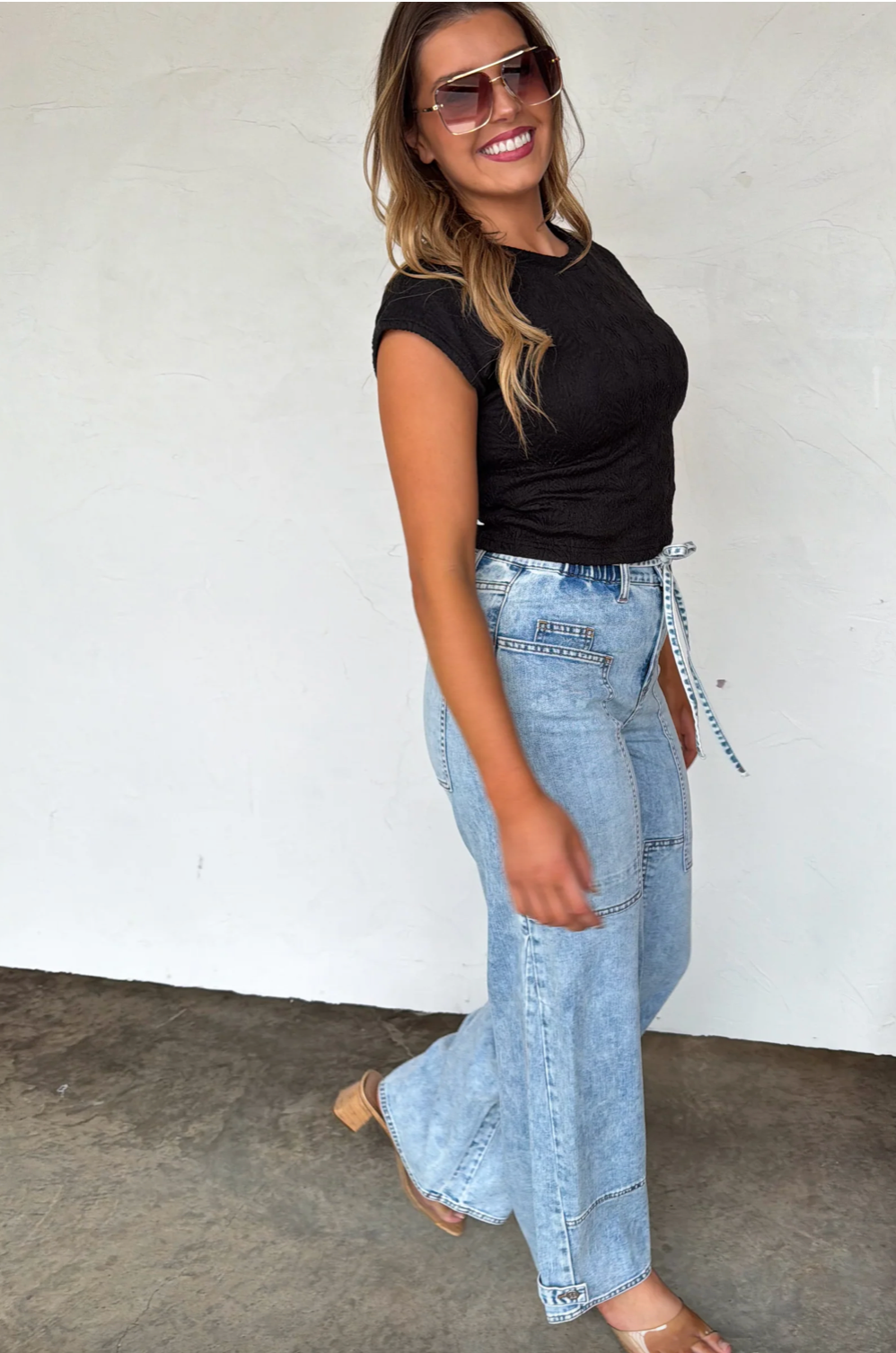 Woman wearing a black top and light blue jeans against a white wall.