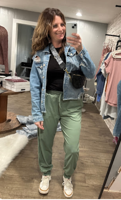 Person taking a mirror selfie wearing a denim jacket, black top, green pants, and white sneakers in a clothing store.