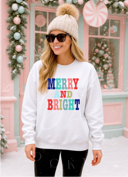 Woman wearing a white sweatshirt with 'Merry and Bright' text, standing in front of a decorated Christmas window.