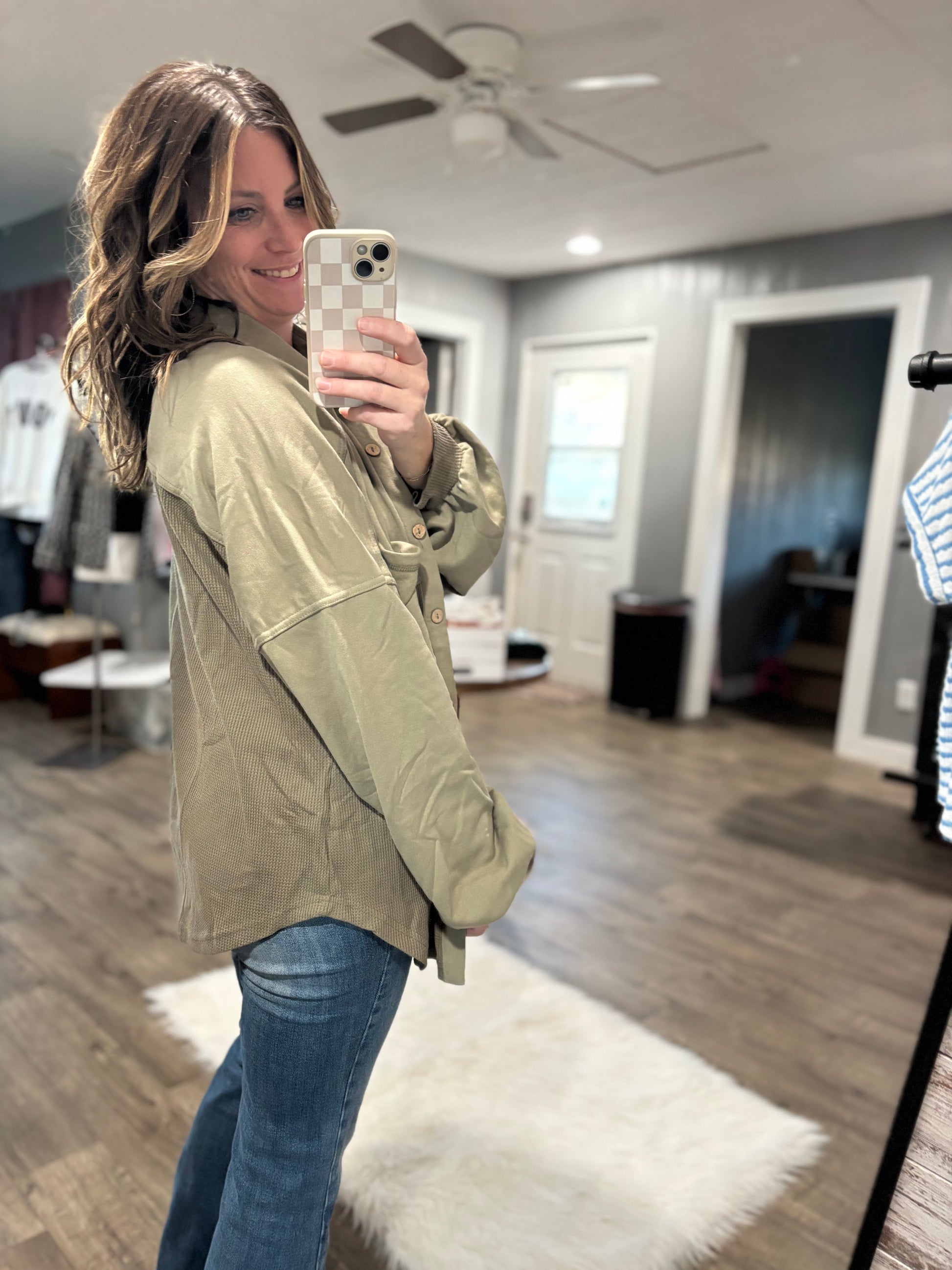 Woman taking a mirror selfie wearing a beige jacket and blue jeans in a room with wooden flooring and a white ceiling fan.