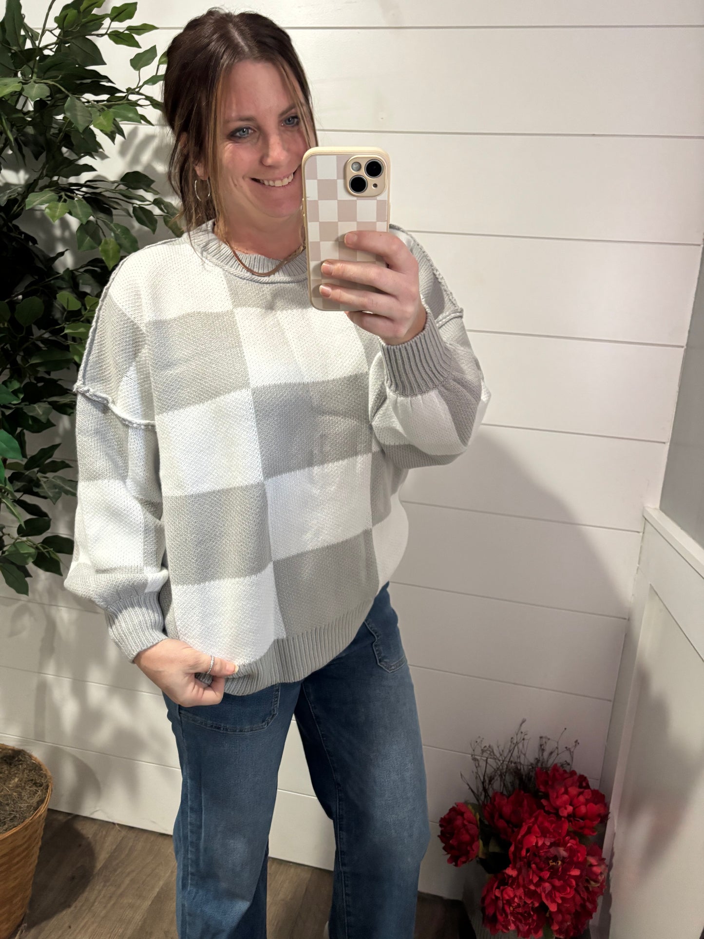 Woman taking a mirror selfie wearing a checkered sweater and jeans, with a white wall and plant in the background.