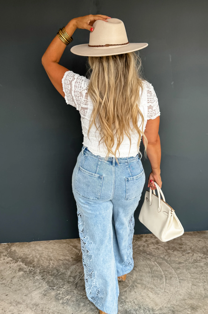 Woman wearing a white lace top, light blue jeans, and a beige hat against a dark gray wall.