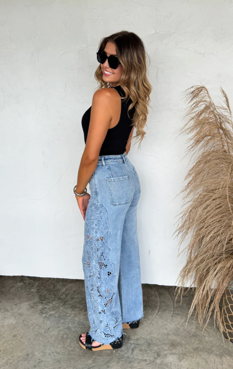 Woman wearing a black top and light blue jeans standing against a white wall with a plant to the side.