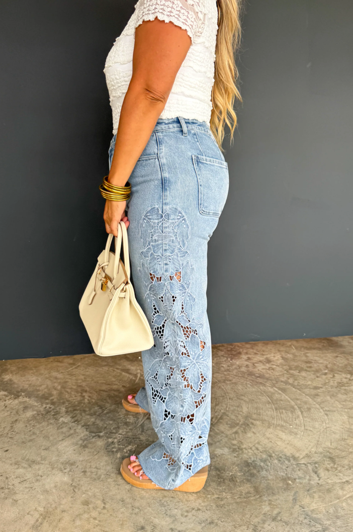 Woman wearing light blue lace pants with a white top and beige handbag against a dark background