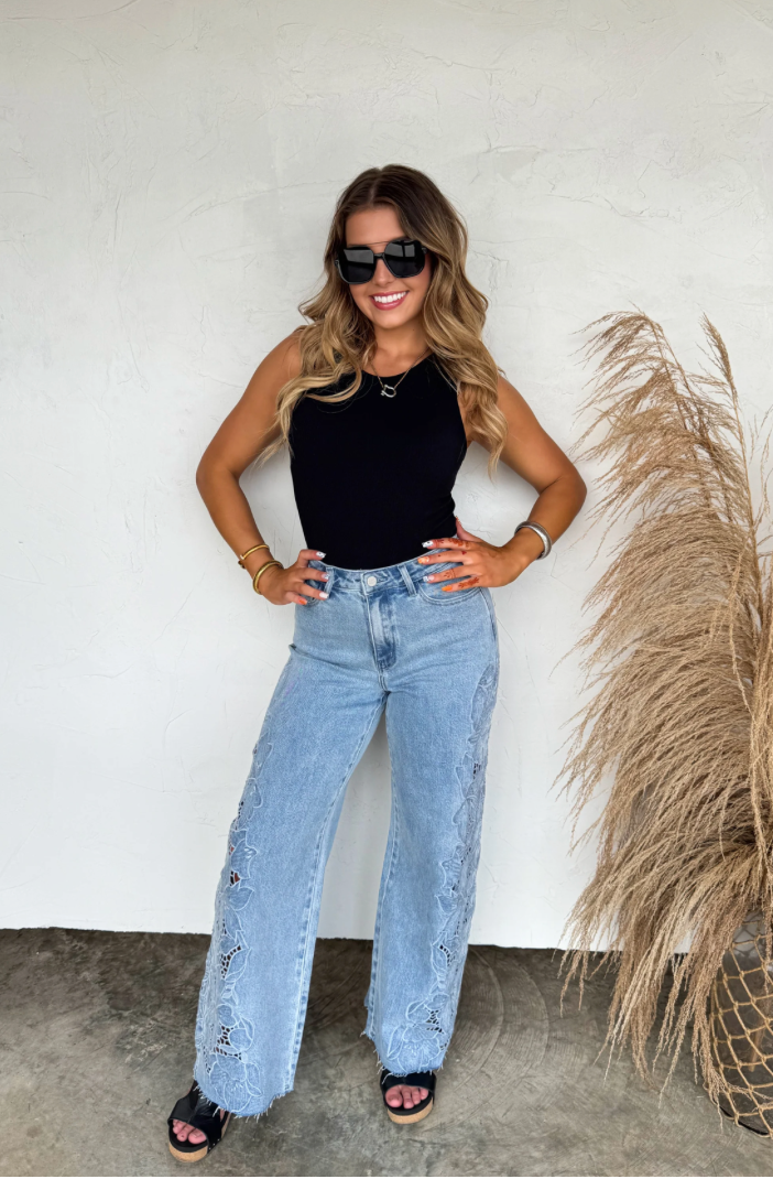 Woman wearing a black top and light blue jeans standing against a white wall with a plant to her right.