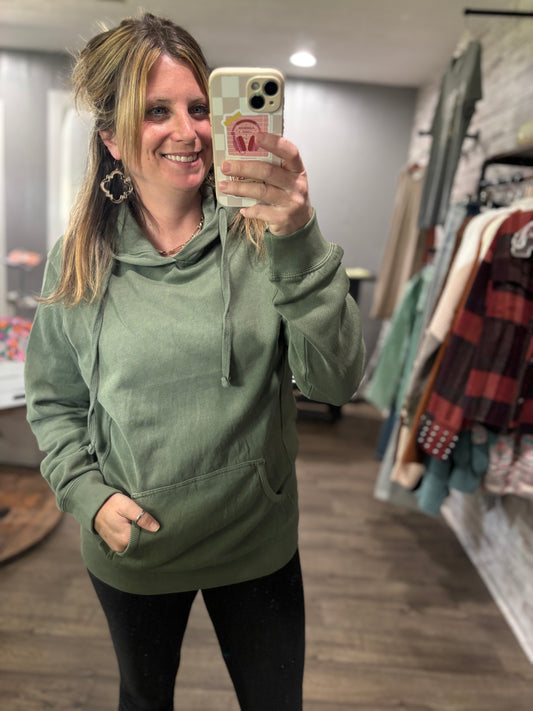 A woman wearing a green hooded sweatshirt with a smile on her face, taking a mirror selfie.
