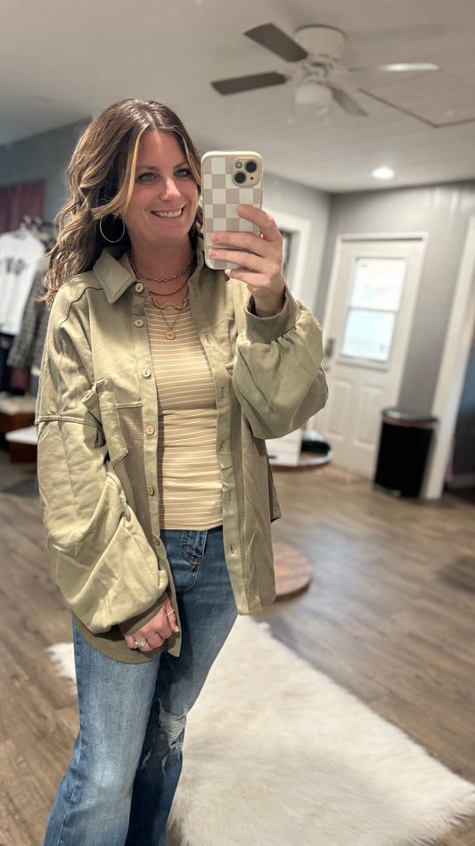 Woman taking a mirror selfie wearing a light green jacket, beige top, and blue jeans in a room with wooden flooring.