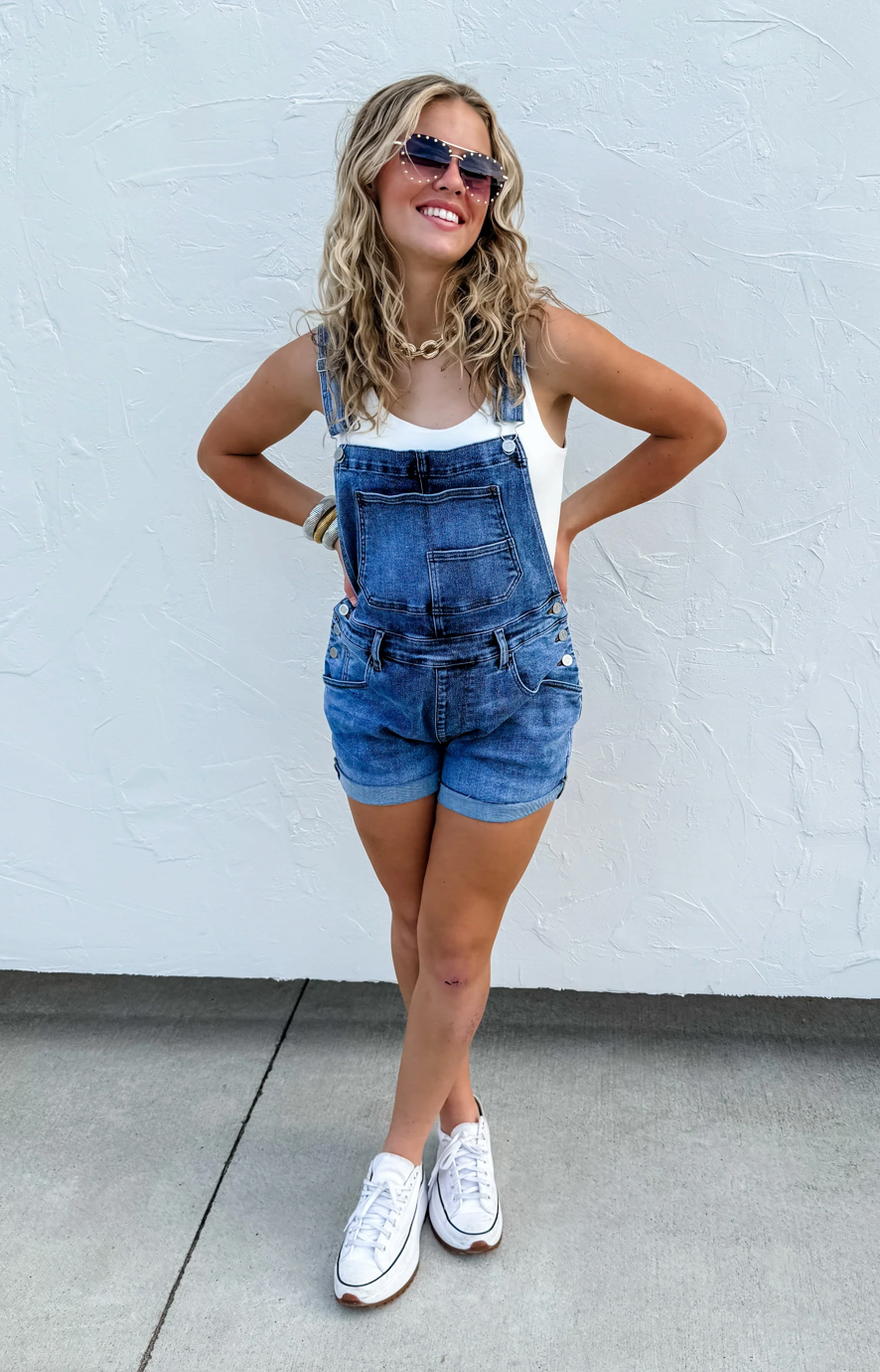 Woman wearing denim overalls and white sneakers against a white wall.